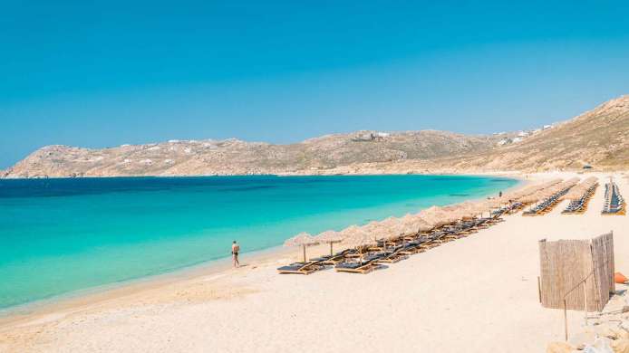 Magnifique plage à l'eau bleue turquoise et au sable blanc. De nombreux bains de soleil sont alignés sur la plage. A faire à Mykonos