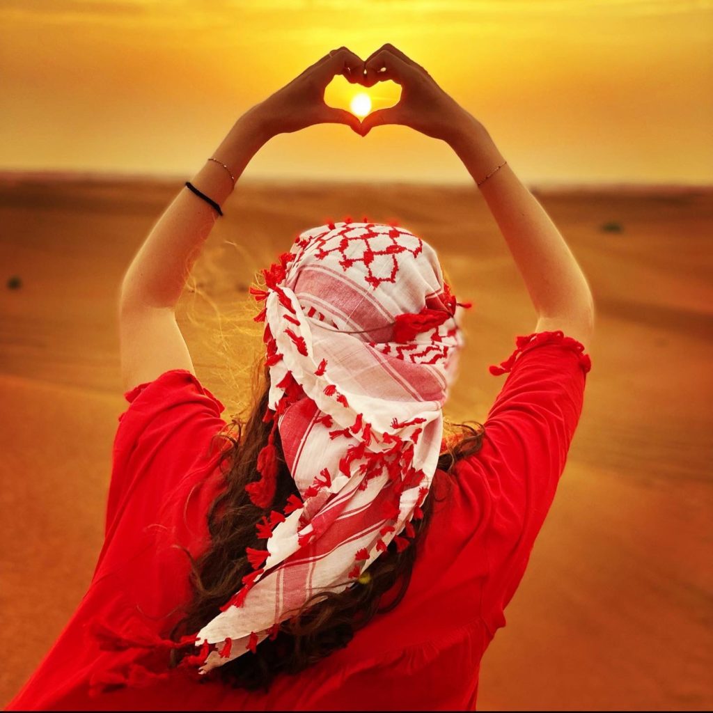 Poto d’une femme de dos qui tiens dans ses mains en forme de cœur le soleil au loin. Le désert est en fond de toile. 