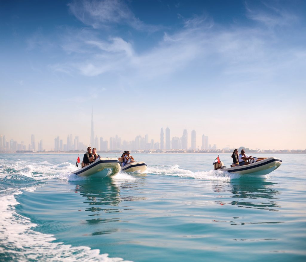 3 petits bateaux à moteur avec deux personnes dans chaque qui découvre la ville par la mer. les buildings sont en fond de toile. Une des activités à faire à Dubai.