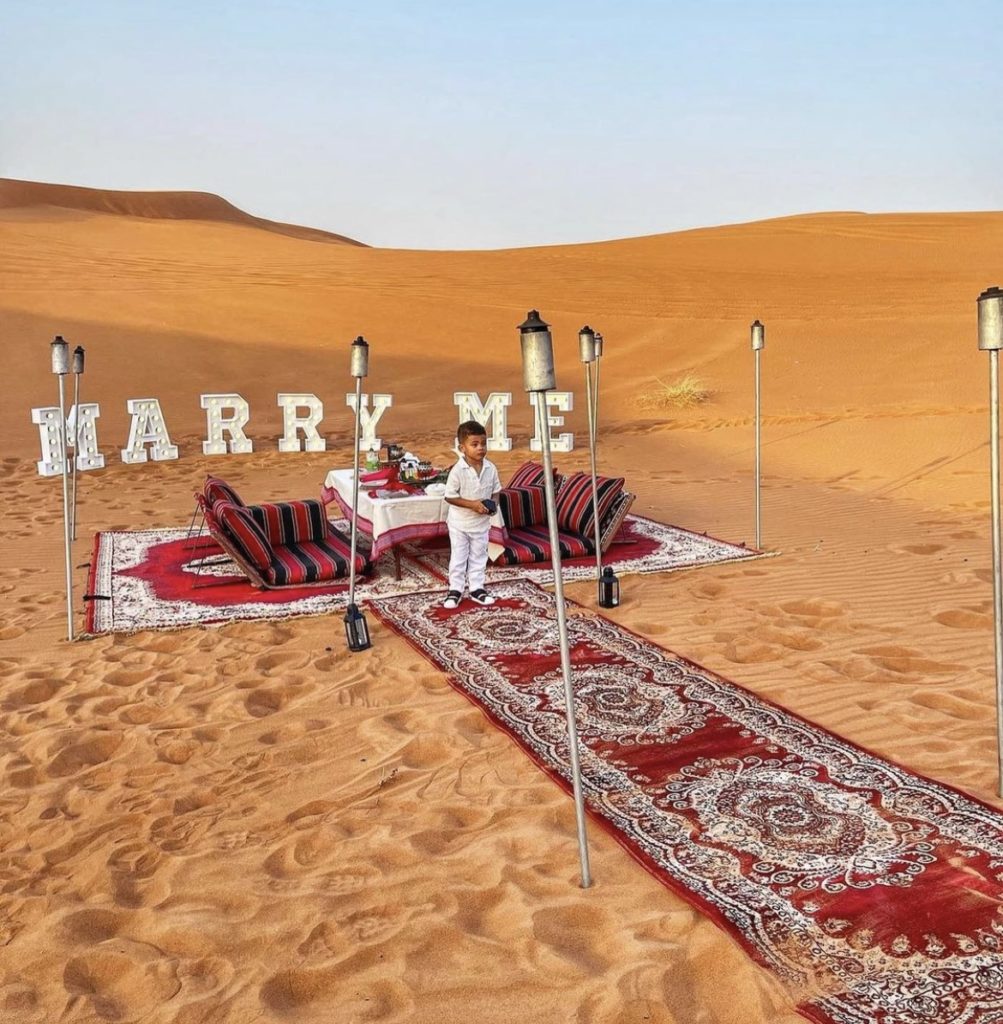 Un tapis rouge qui mène à deux fauteuils en plein milieu du désert pour une future demande en mariage. Un enfant se tient au centre avec les alliances.