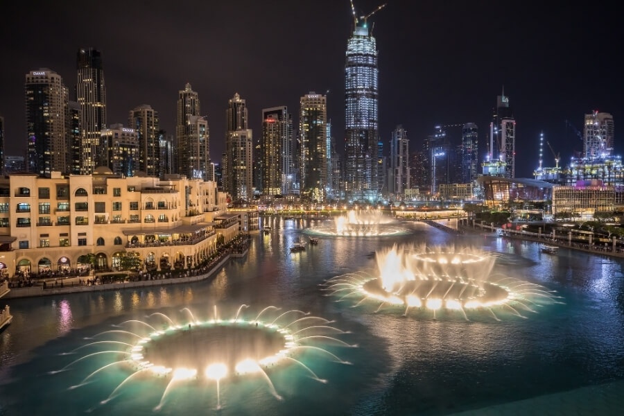 Spectacle des fontaines éclairées avec vue sur toute la place principale située aux pieds de la Burj Khalifa à Dubai. Les buildings sont tous éclairés.